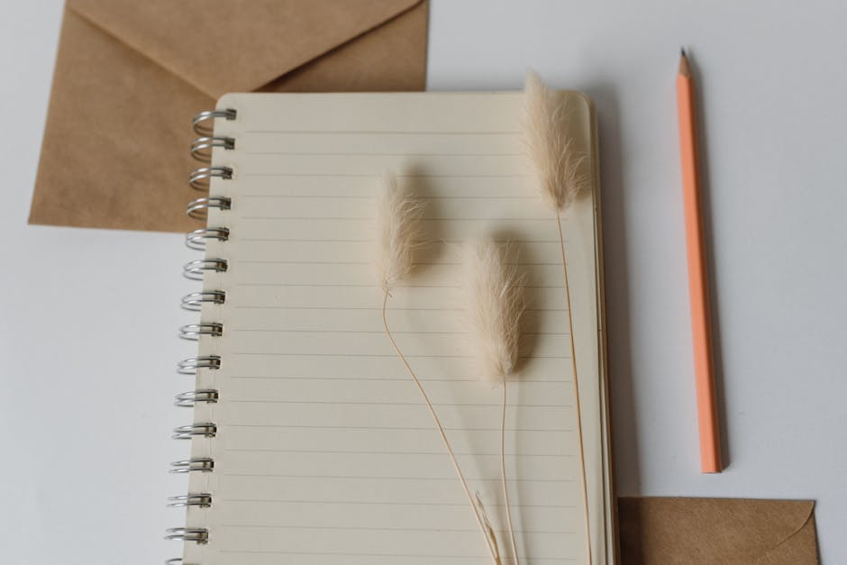 Top view of a notebook with envelopes, pencil, and soft decorative grass on a white surface.